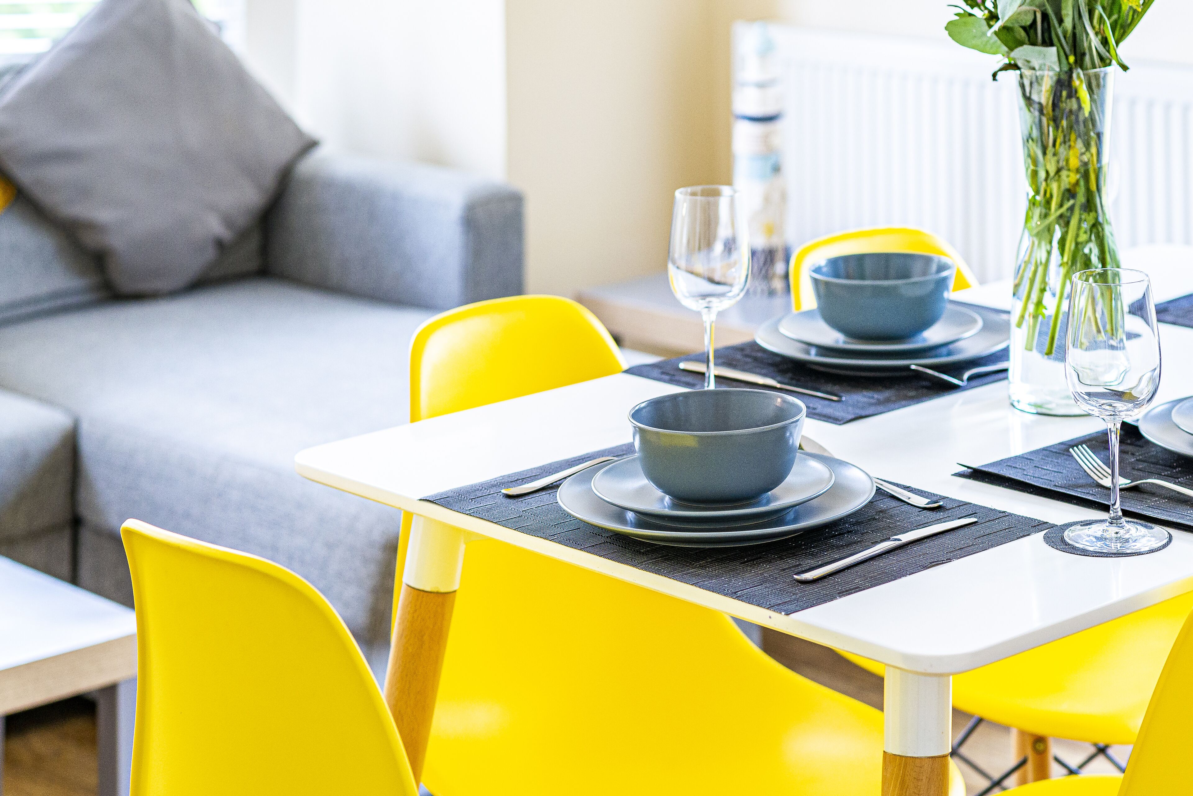 Un coin repas avec des chaises jaunes, une table blanche et un canapé gris en arrière-plan.