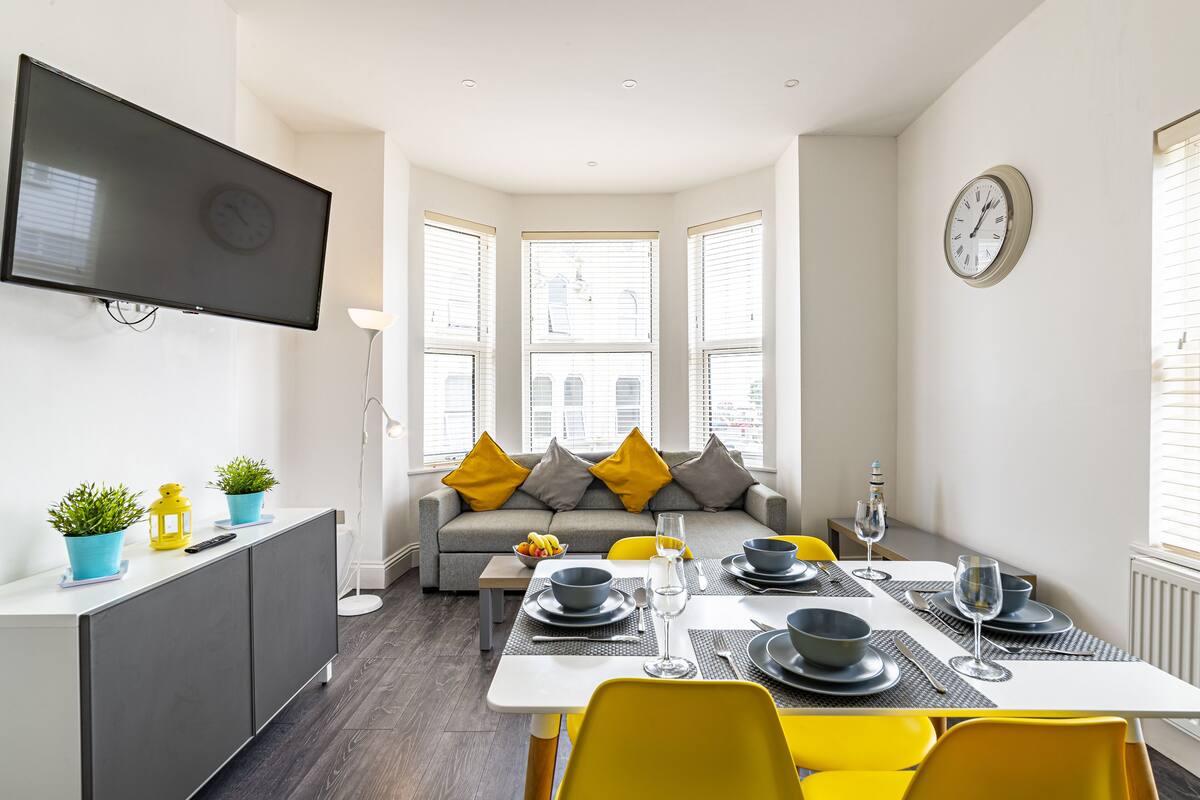 Ein moderner Essbereich mit einem weißen Tisch, gedeckt für vier Personen, gelben Stühlen und einem grauen Sofa mit gelben Kissen.