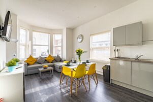 A modern kitchen with a built-in oven, a dining area with yellow chairs, and a grey sofa with yellow cushions.