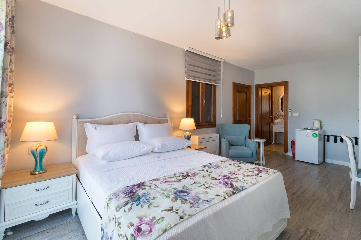 A hotel room with a bed, two bedside tables with lamps, a chair, a desk, and a refrigerator.
