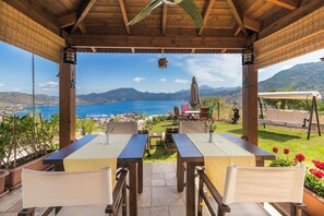 A covered outdoor seating area with tables and chairs, overlooking a scenic view of a body of water and mountains. - Harmanyeri Tatilevi Selimiye (Marmaris)