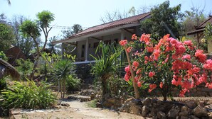 A house with a stone foundation, a porch, and a garden with various plants and a flowering tree. - Join Homestay (Kuta)