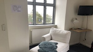 A small, white, single-bed room with a window, a radiator, a desk with a lamp, and a TV mounted on the wall. - Hotel Crone (Grenaa)