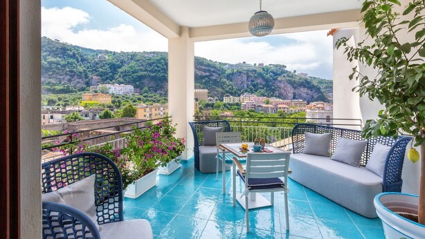Un balcón con vistas a un pueblo en la ladera, una mesa con sillas y una lámpara colgante.