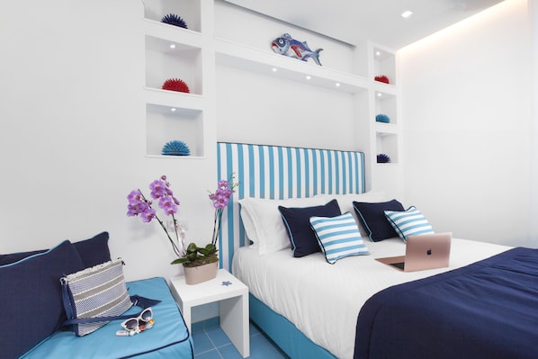 Une chambre moderne avec une tête de lit à rayures, un lit avec une literie bleu marine et blanche, un ordinateur portable posé sur le lit, une petite table de chevet blanche avec une plante et des lunettes de soleil, et des poissons décoratifs sur des étagères.