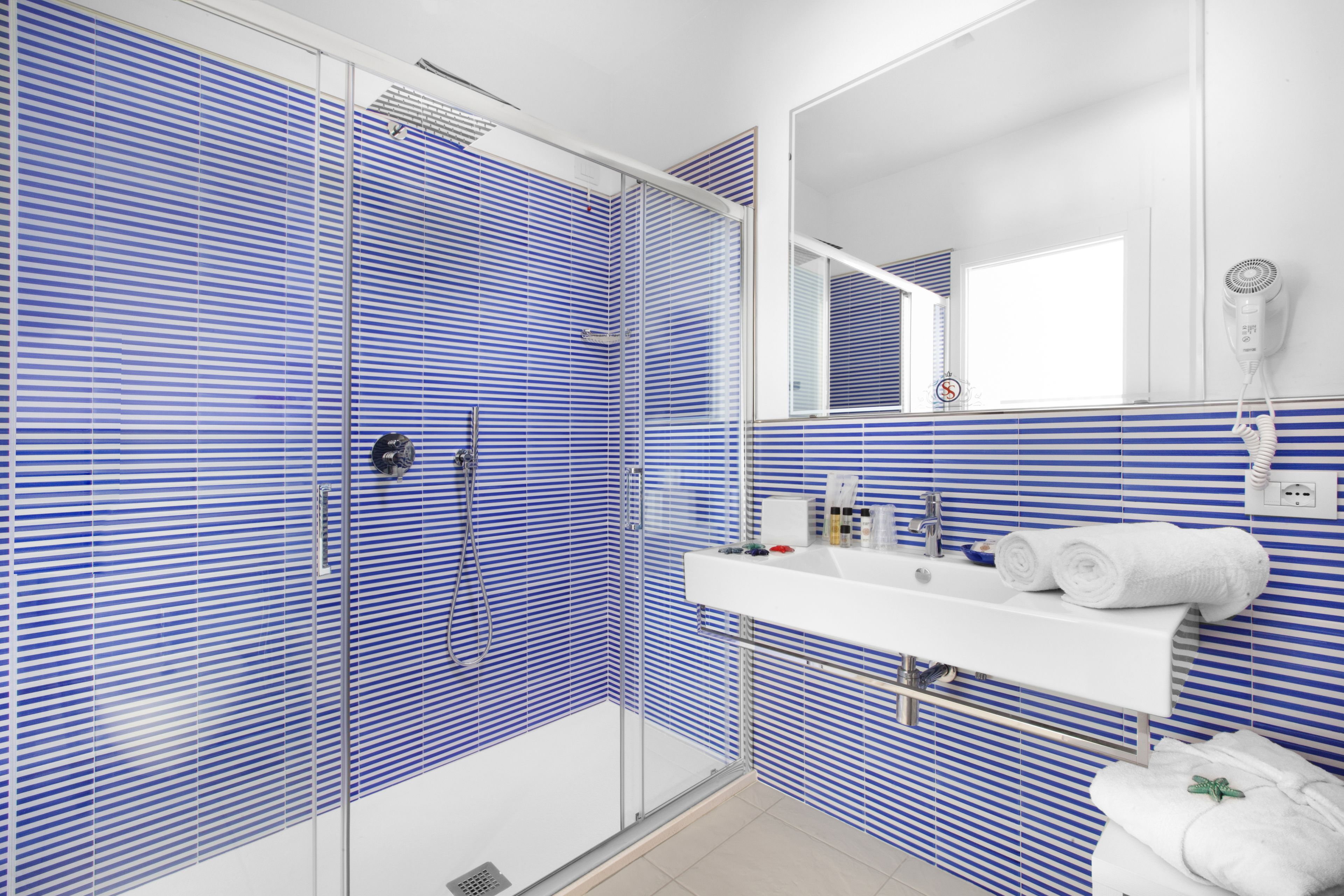 A modern bathroom with blue-striped tiles, a glass-enclosed shower, a white sink with a mirror, and a hairdryer on the wall.