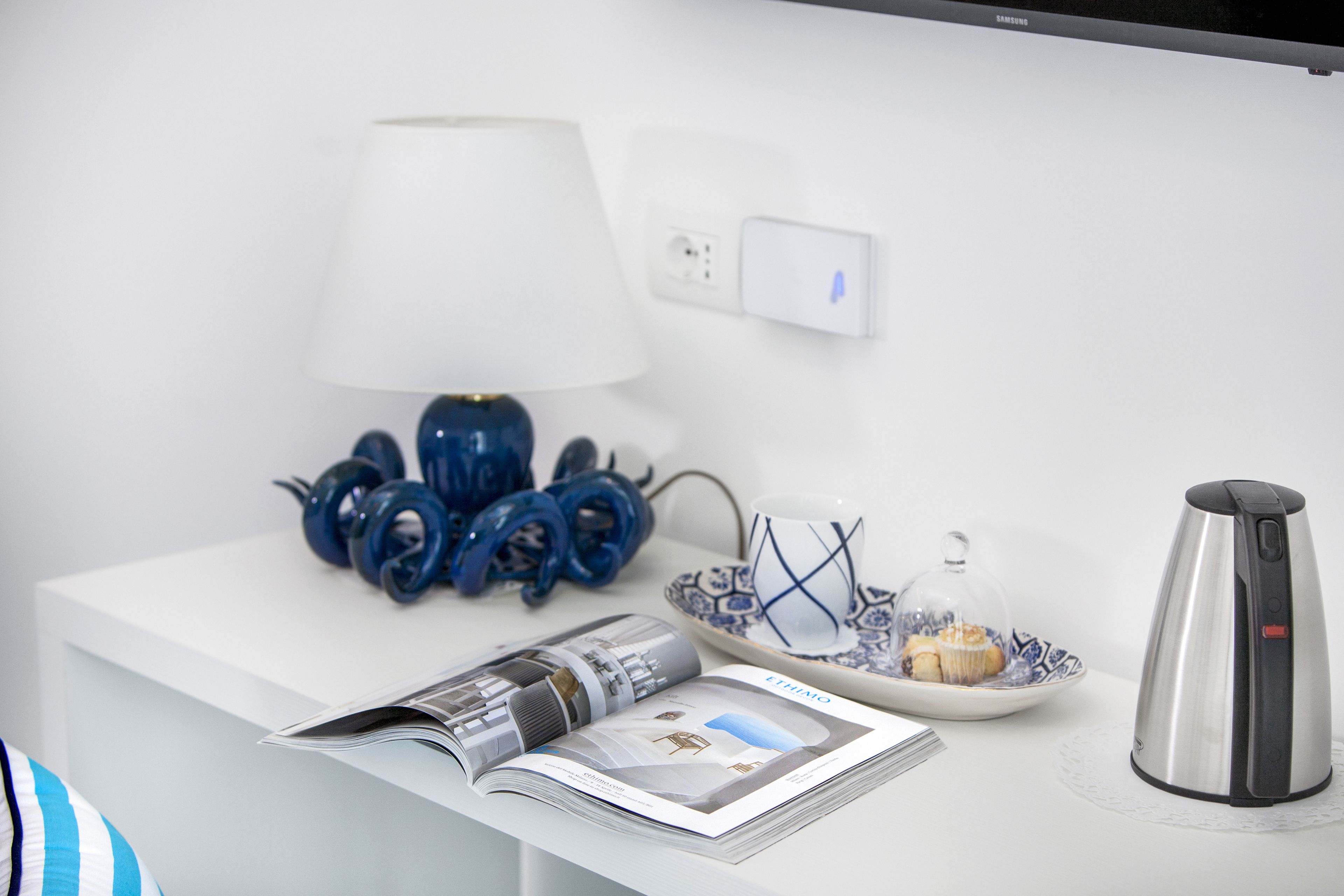 Une table blanche avec une lampe, une assiette contenant un muffin, une bouilloire et un magazine.