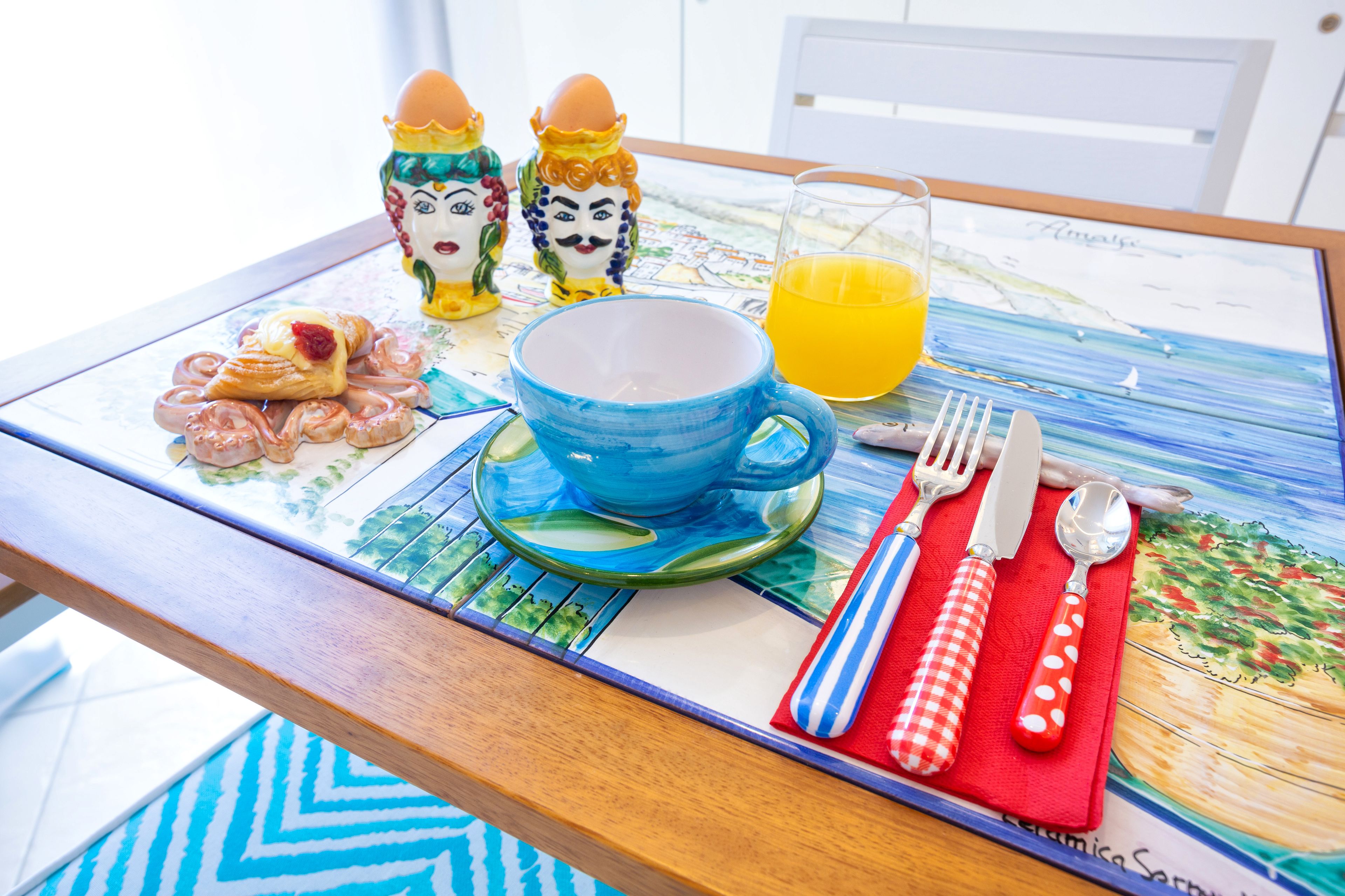 Et morgenbord med en blå kop, appelsinjuice, croissanter, salt- og peberkværn og en serviet i en rød-hvid ternet servietholder.