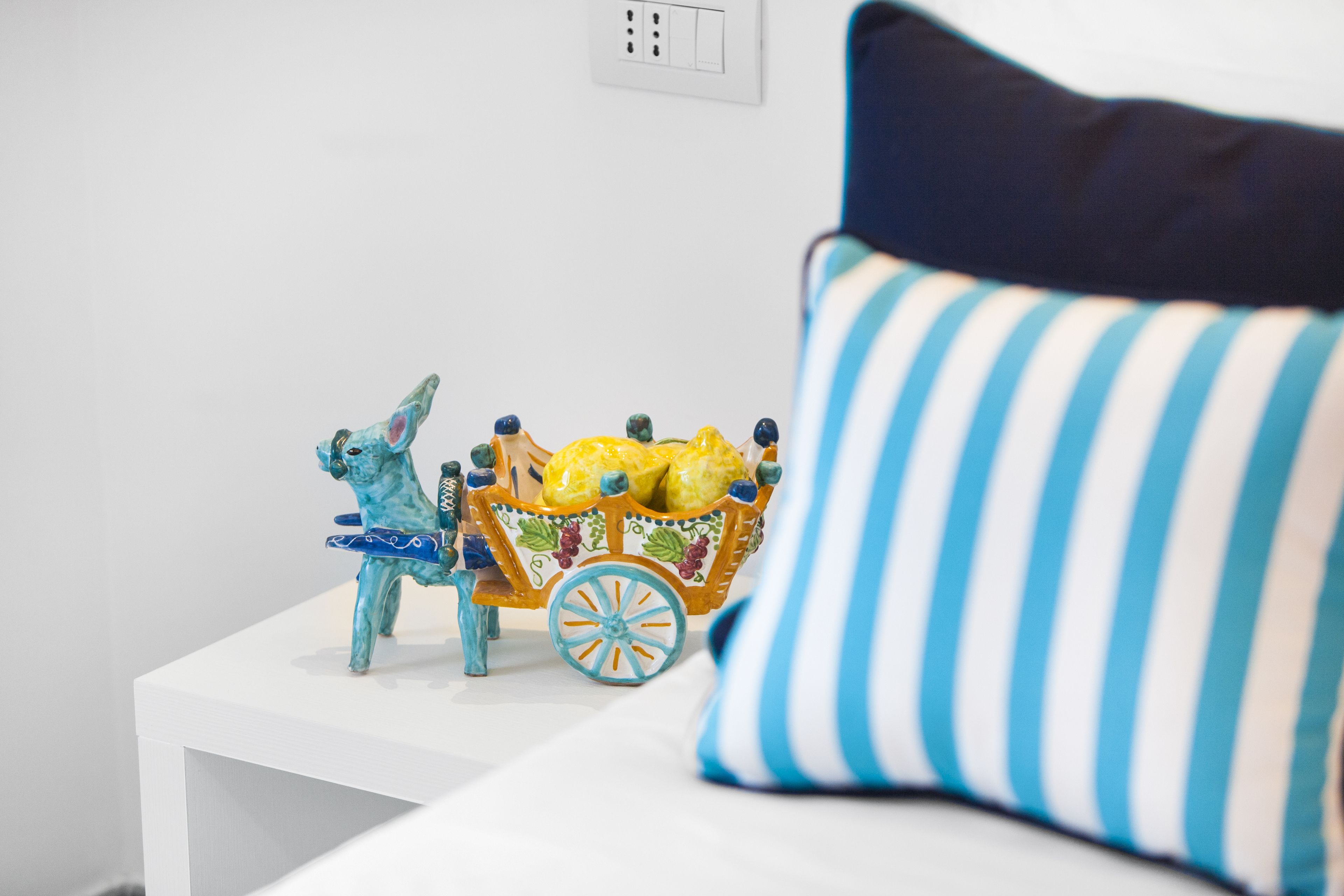 A white bedside table with a decorative ceramic horse and carriage, a striped pillow, and a white wall with a power outlet.