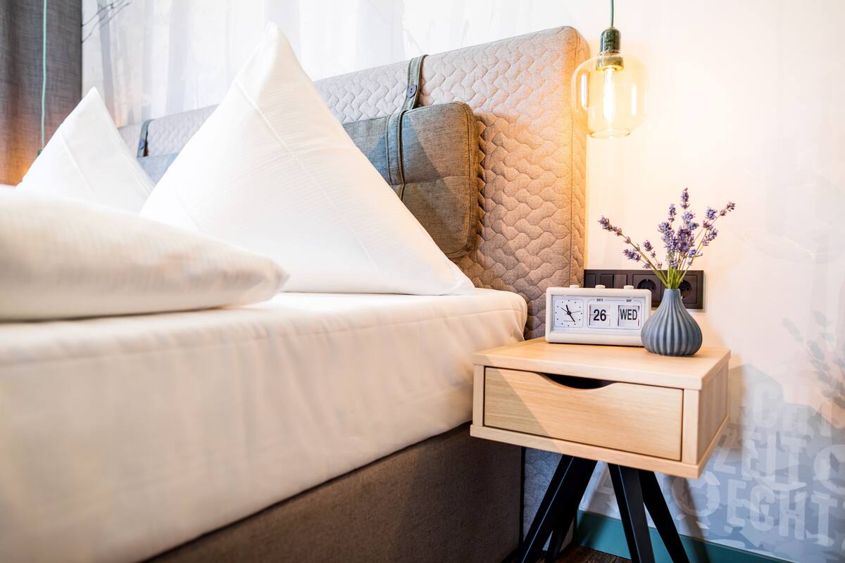 Una cama bien hecha con ropa blanca, una mesita de noche con un reloj y un jarrón de flores, y una pared con diseño texturizado.