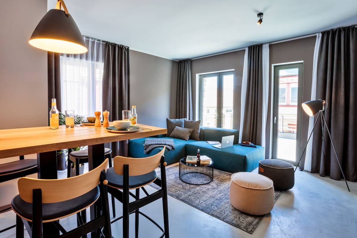 Una sala moderna con una mesa de comedor de madera, dos sillas, un sofá azul, una pequeña mesa redonda de centro y una lámpara de pie.