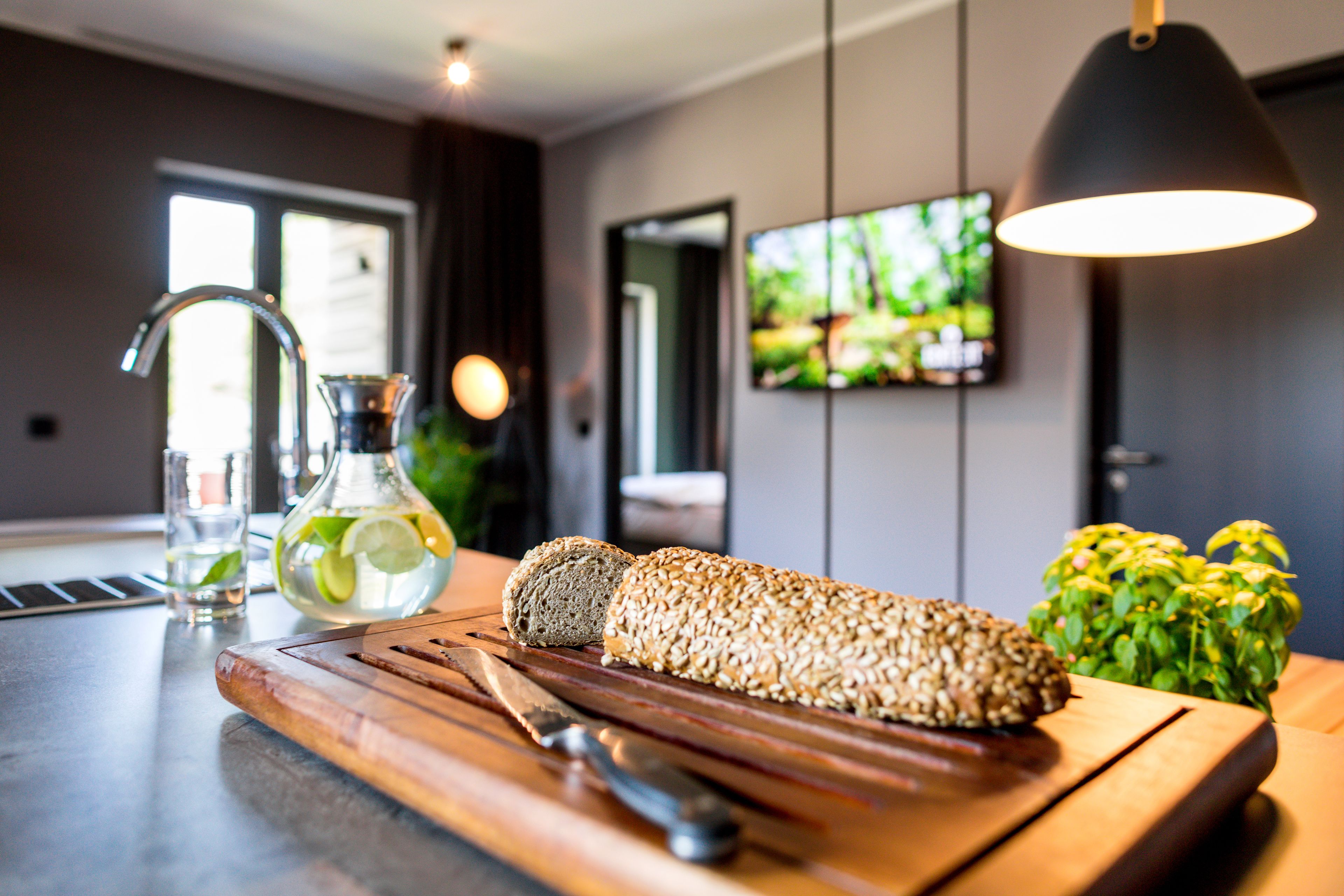 A kitchen counter with a loaf of bread, a knife, a cutting board, a glass of water, and a bottle of lime.