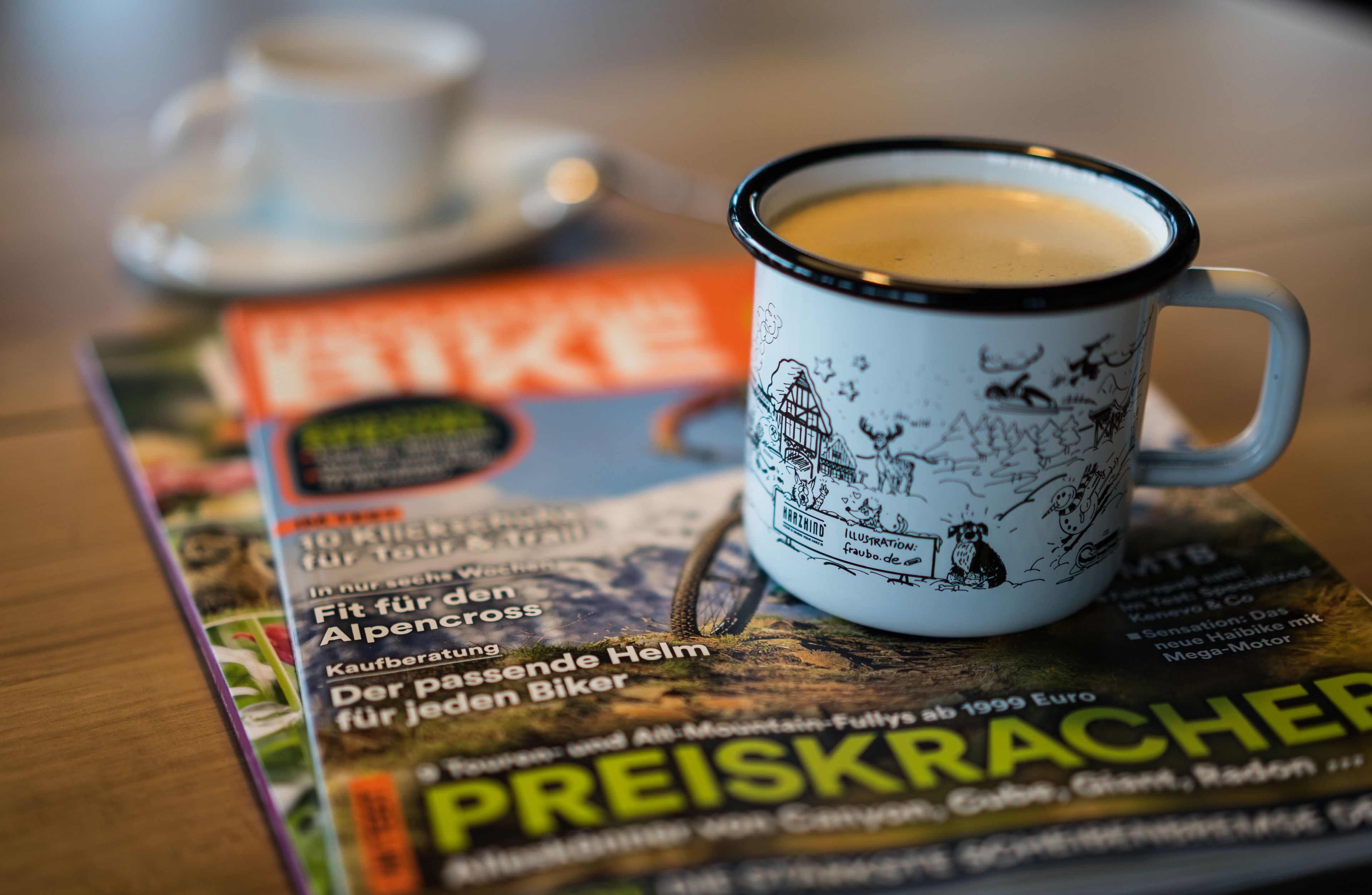 Una taza de café con diseño de bicicleta sobre una mesa, junto a una revista con texto en alemán.