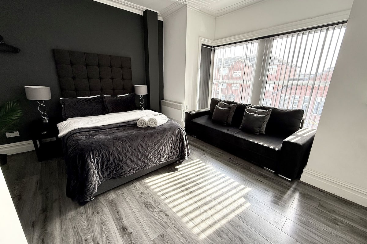 A modern bedroom with a large bed, a dark headboard, a bedside table with lamps, a black sofa, and a large window with blinds.