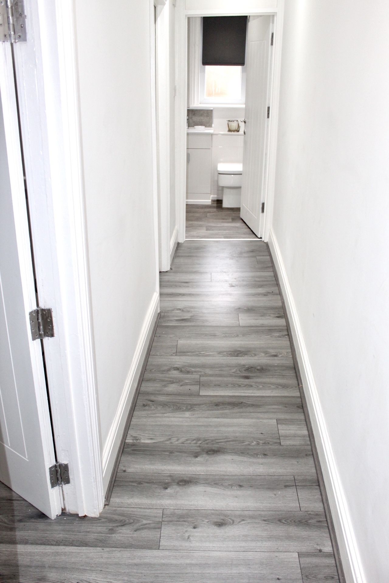 A narrow hallway with a wooden floor and white walls, leading to a bathroom with a toilet and sink.