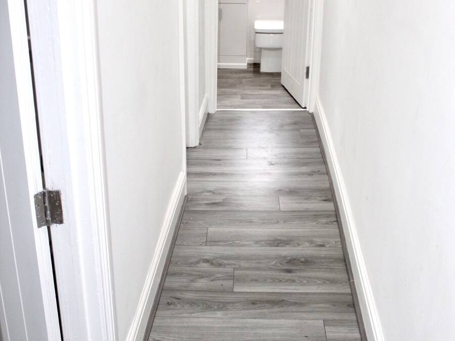 A narrow hallway with a wooden floor and white walls, leading to a bathroom with a toilet and sink.