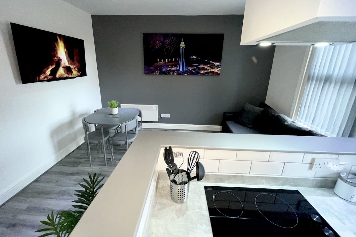 A modern kitchen with a built-in stove, a dining area with a small table and chairs, and a large flat-screen TV mounted on the wall.