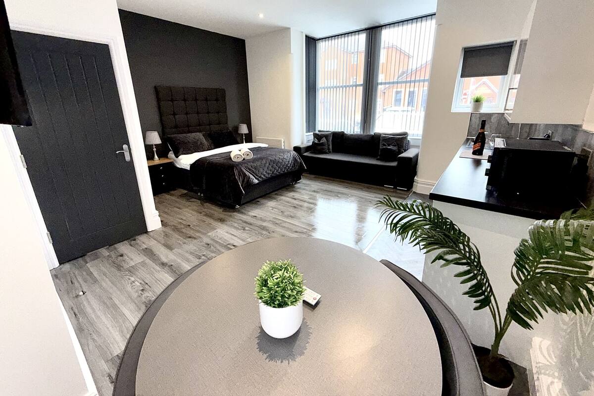 A modern bedroom with a bed, bedside tables, a sofa, a small kitchen area, and a dining table with a potted plant.