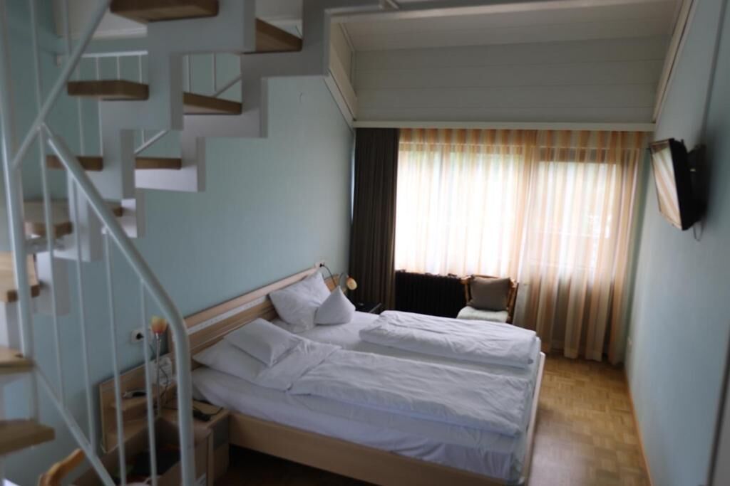 A bedroom with two beds, a wooden staircase, and a view of the outdoors through curtains.