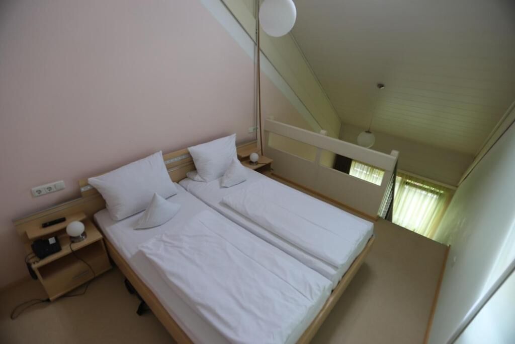 A single-bed room with a wooden headboard, white bedding, a bedside table with a lamp, and a window with curtains.
