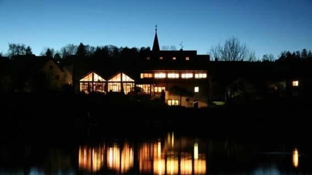 A building with a prominent tower and multiple windows reflecting on a calm body of water. - Pension Krämerhof Kulz (Thanstein)