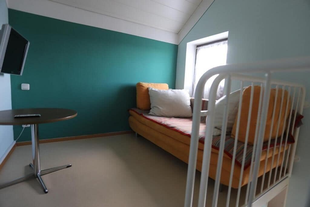 Una habitación con paredes color turquesa, una cama con ropa de cama blanca y naranja, una mesita redonda y un televisor montado en la pared.