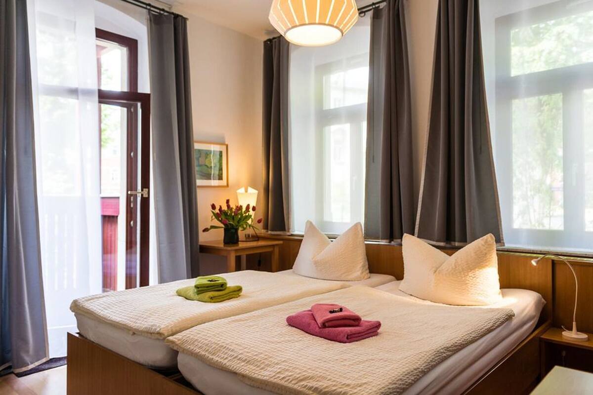 Habitación de hotel con dos camas, una mesita de noche con lámpara, una ventana con cortinas y una puerta que da al exterior.