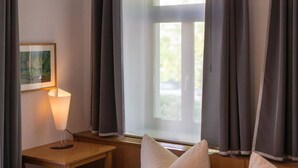 Ein Zimmer mit Fenster, dunklen Vorhängen, einer Lampe auf einem Holztisch und einem gerahmten Bild an der Wand.