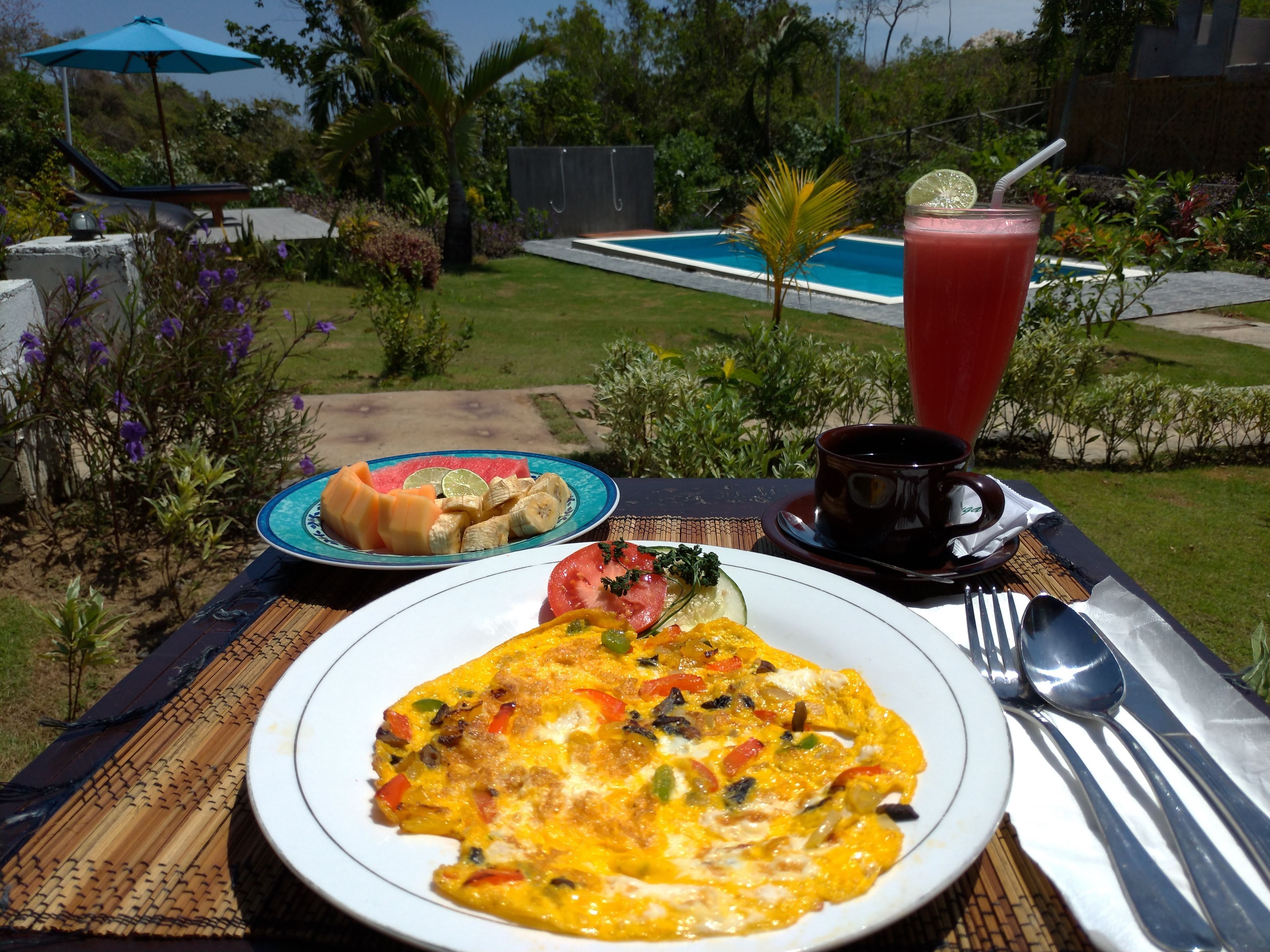 Ein Frühstückstisch im Freien mit Eiern, Obst, einer Kaffeetasse und einem Glas Saft, mit Blick auf einen Pool und einen Garten.