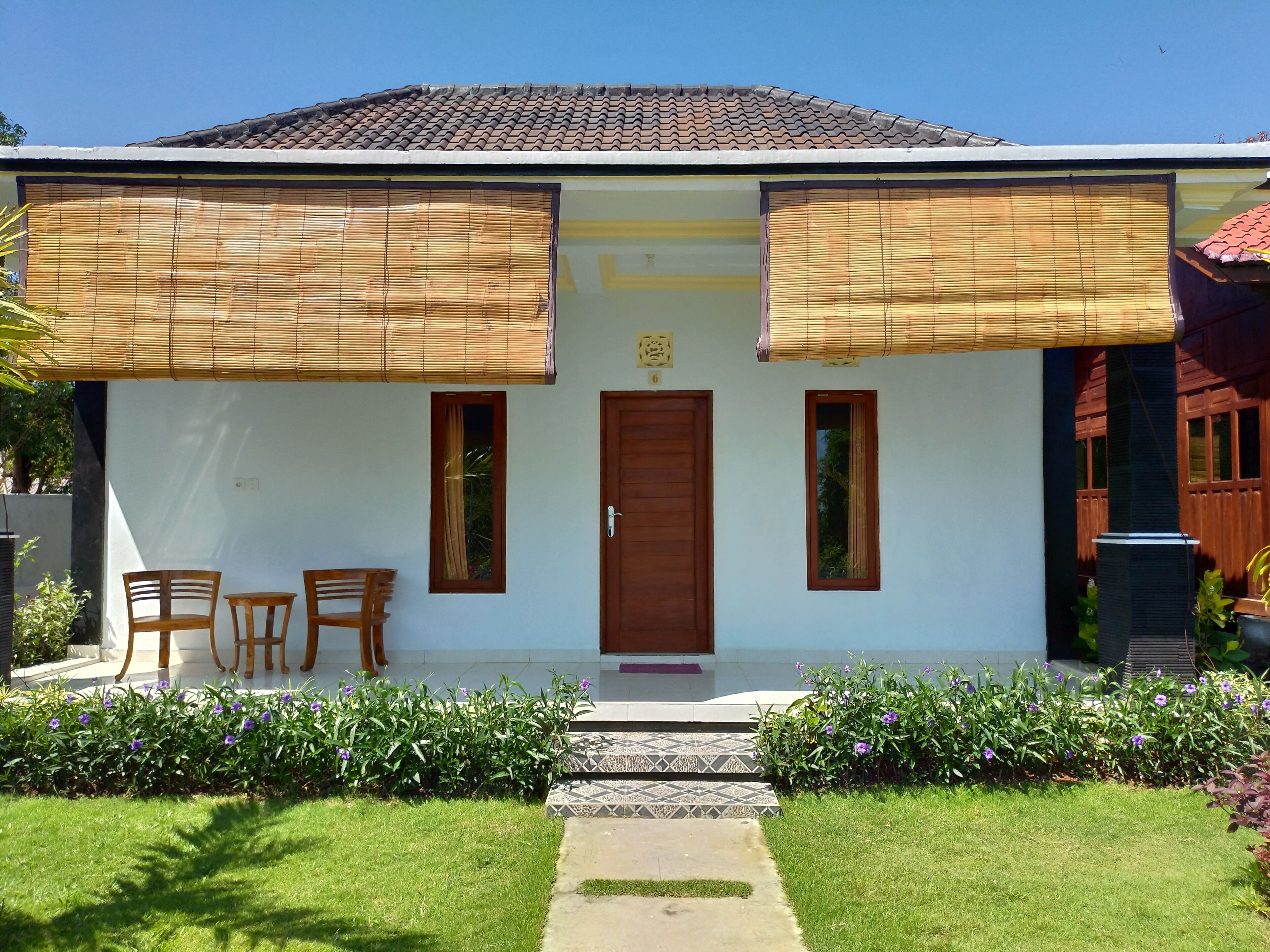 A house with a tiled roof, a wooden door, and two windows with curtains. There are two wooden chairs and a small table on the porch.