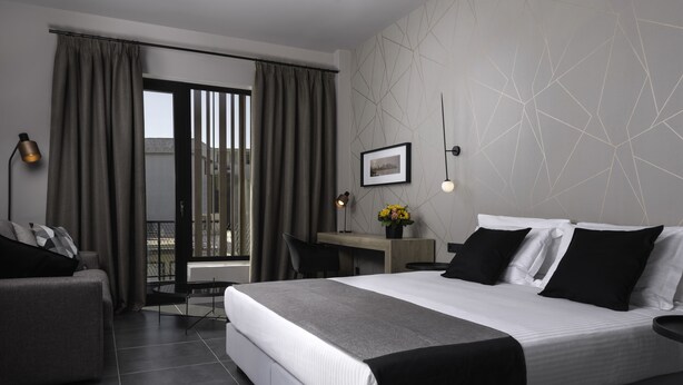 Quarto de hotel moderno com uma cama grande, uma escrivaninha, uma cadeira e vista para o exterior através de uma porta deslizante.