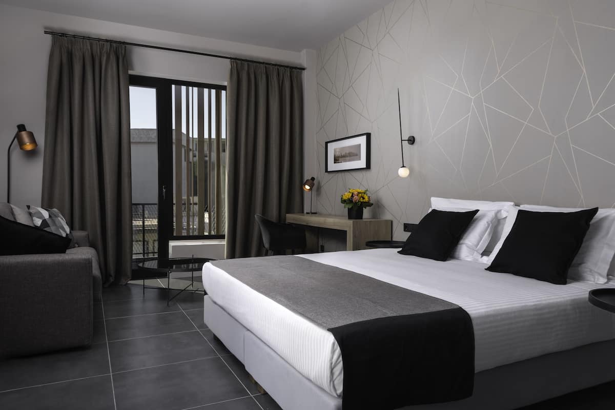 A modern hotel room with a large bed, a desk, a chair, and a view of the outdoors through a sliding door.