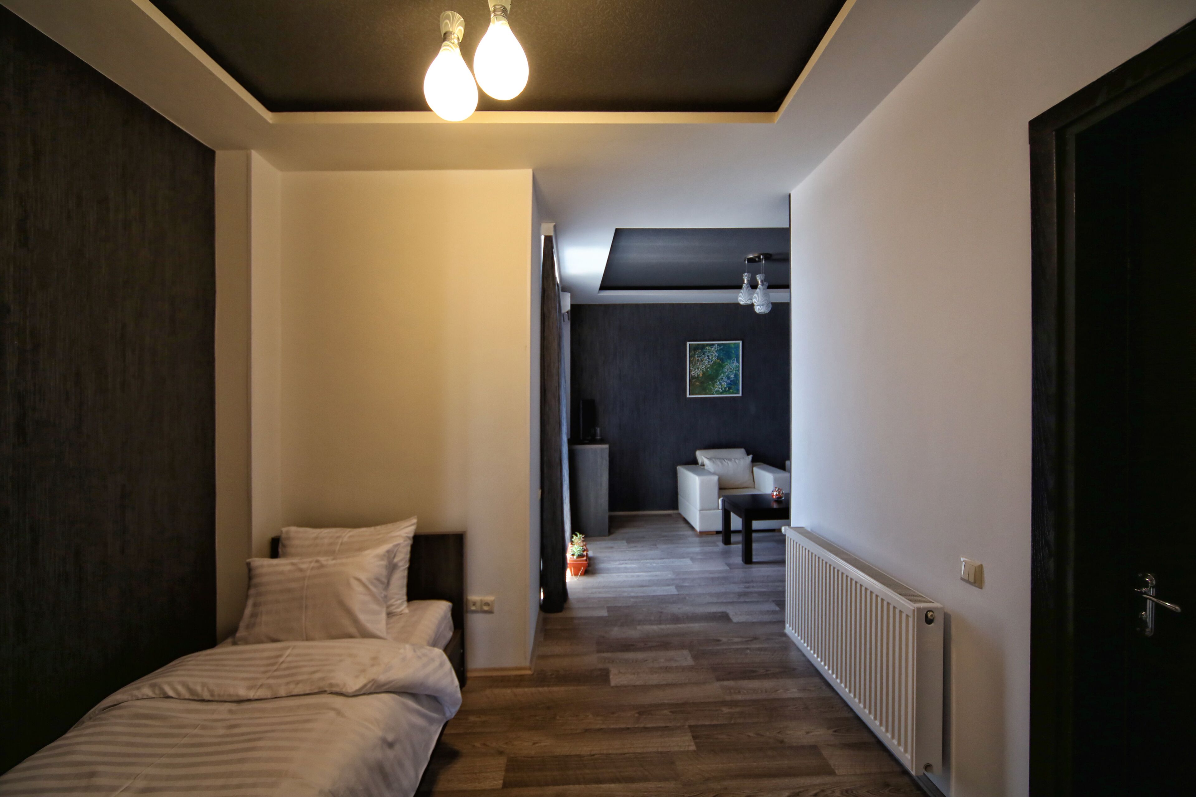 Una habitación de hotel moderna con suelo de madera, una cama con ropa de cama blanca, una mesita y una silla. Se observa un radiador a lo largo de la pared y una puerta de color oscuro a la derecha.