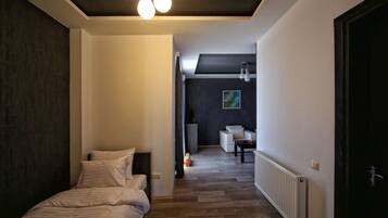 Una habitación de hotel moderna con suelo de madera, una cama con ropa de cama blanca, una mesita y una silla. Se observa un radiador a lo largo de la pared y una puerta de color oscuro a la derecha.