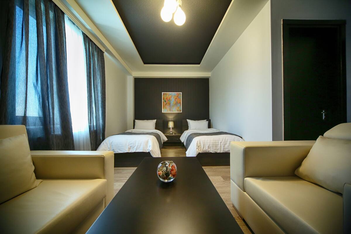 A hotel room with two beds, a sofa, a coffee table with a decorative bowl, and a painting on the wall.