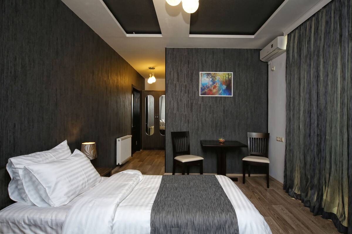 A modern hotel room with a large bed, a desk, two chairs, and a painting on the wall.