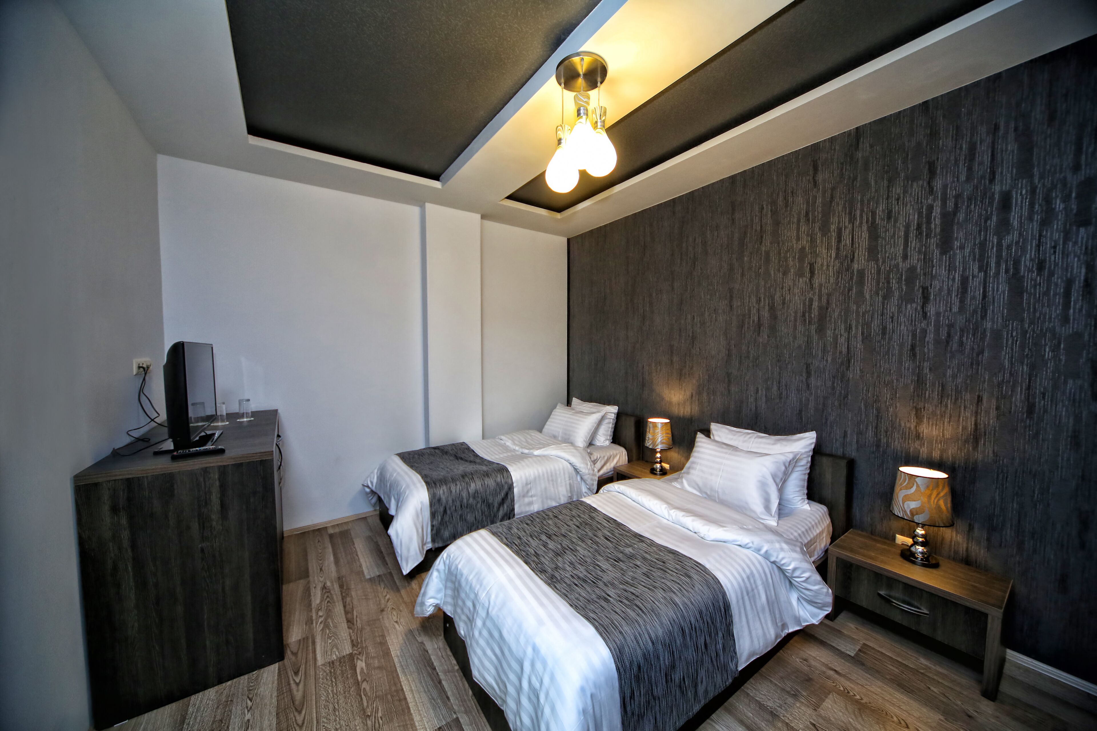 A modern hotel room with two beds, a wooden dresser, a flat-screen TV, and a ceiling light fixture.