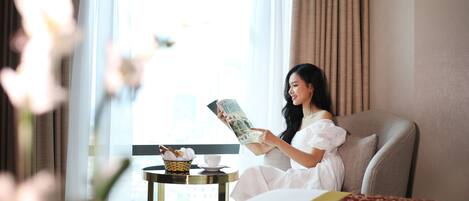 A woman sitting on a bed in a hotel room, reading a magazine. There is a small table with a basket of pastries and a cup.