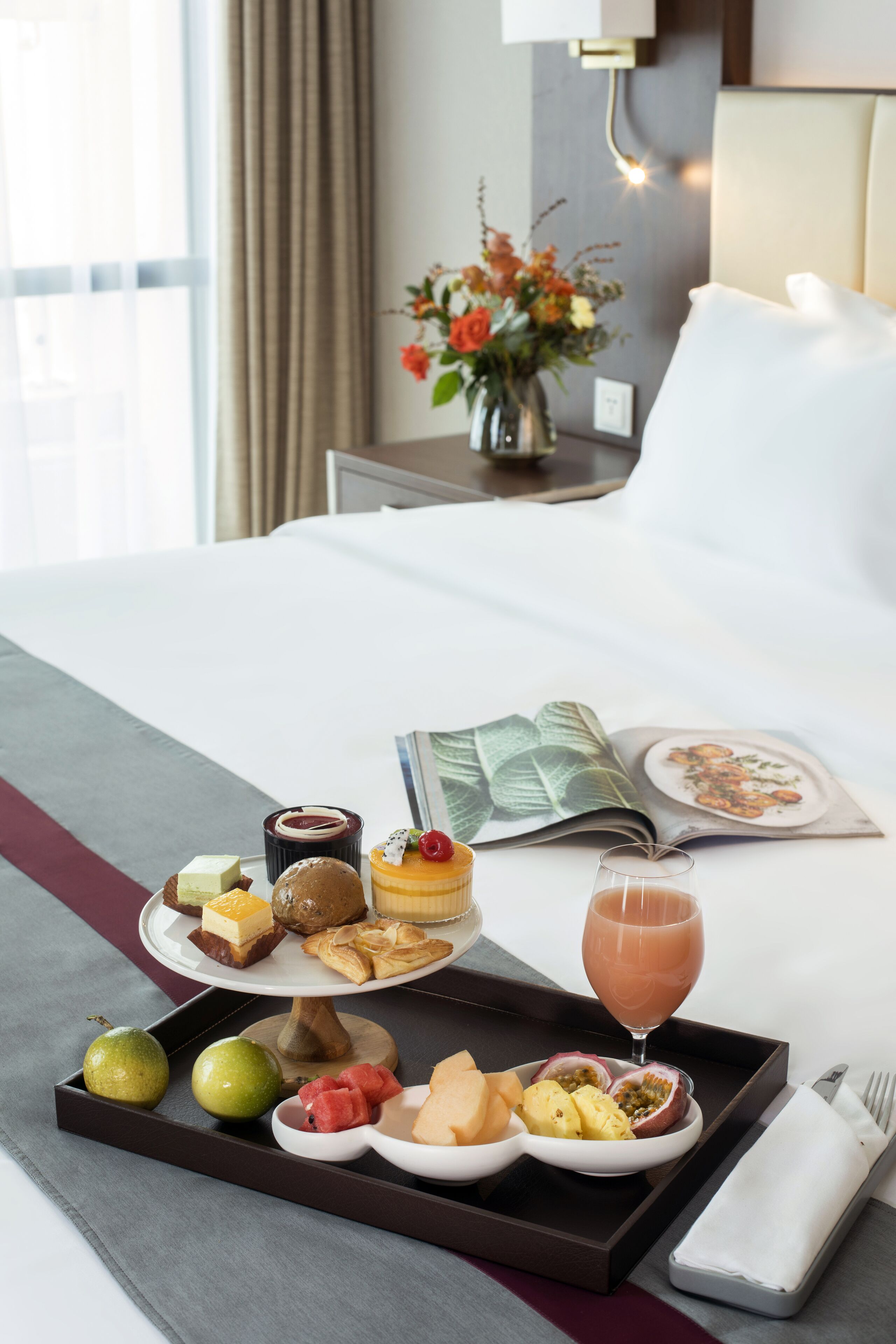 A neatly arranged breakfast tray with various fruits, pastries, and a drink on a hotel bed.