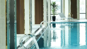 An indoor swimming pool with a clear water surface, a lounge chair, a small table with a potted plant, and a window with a view of buildings. - Grand Vista Hanoi (Hanoi)