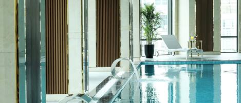 Una piscina interior con superficie de agua cristalina, una silla de descanso, una mesita con una planta en maceta y una ventana con vistas a edificios.