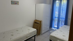 A small, clean room with two single beds, a wooden chair, a white wardrobe, and a blue curtain covering a window.
