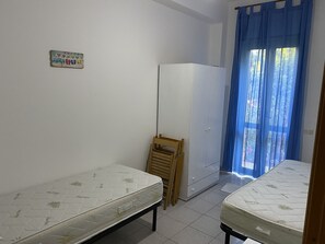 Una stanza piccola e pulita con due letti singoli, una sedia di legno, un armadio bianco e una tenda blu che copre una finestra.