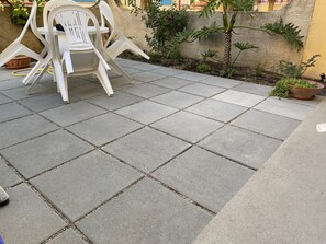 Um espaço de pátio com azulejos de pedra quadrados, cadeiras de plástico branco e uma pequena palmeira.