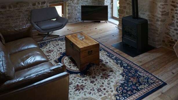 Une pièce de style « loft », avec des poutres en bois apparentes, un mur en pierre, une cheminée, un canapé en cuir, une malle en bois et un tapis à motifs.