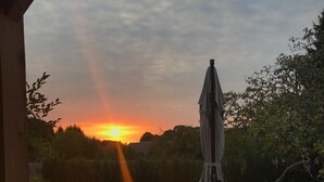 Une vue sur le coucher de soleil, prise depuis une fenêtre donnant sur un jardin où se trouvent un parasol et une petite table avec un saladier.