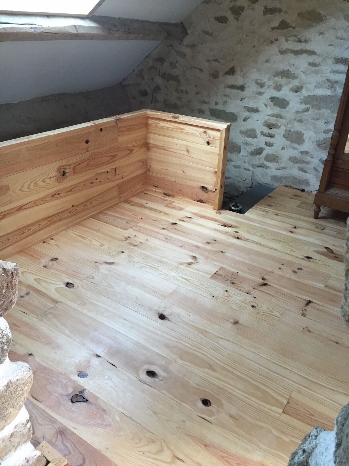 A room with a sloped ceiling, wooden flooring, and a stone wall.