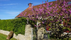 Un mur de pierre avec un portail en métal, un pilier de pierre et un bâtiment doté d’un toit recouvert de tuiles rouges et agrémenté d’arbustes à fleurs violettes.