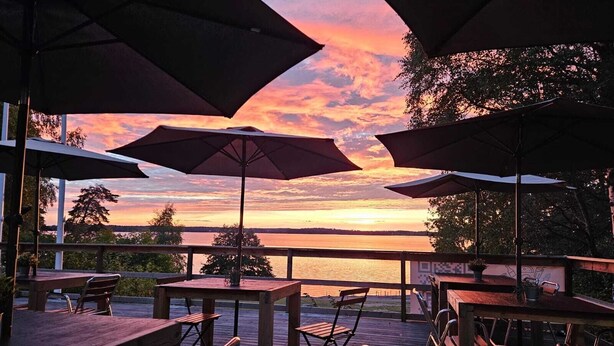 An outdoor seating area with tables and chairs, umbrellas, and a view of a sunset over a body of water. - Kallhällsbaden Vandrarhem - Hostel (Jarfalla)