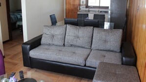 A living room with a grey sofa, a coffee table, and a TV. There is a kitchen area with a refrigerator and a window. - New apartment, 1A center of Villarrica (Villarrica, Araucania)